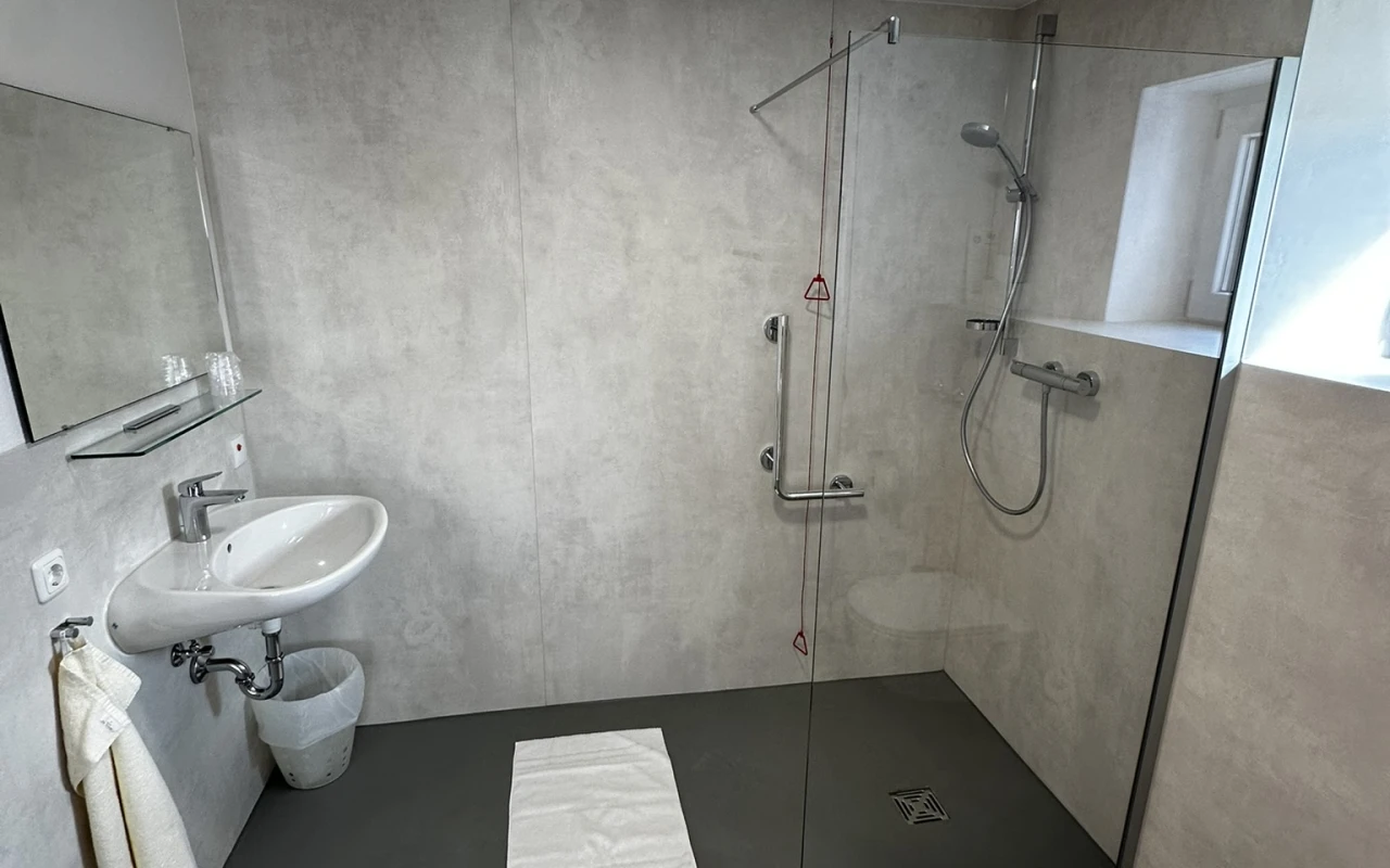 Bild Barrierefreies Badezimmer mit bodengleicher Dusche, Haltegriffen, Duschsitz, Waschbecken und WC.
